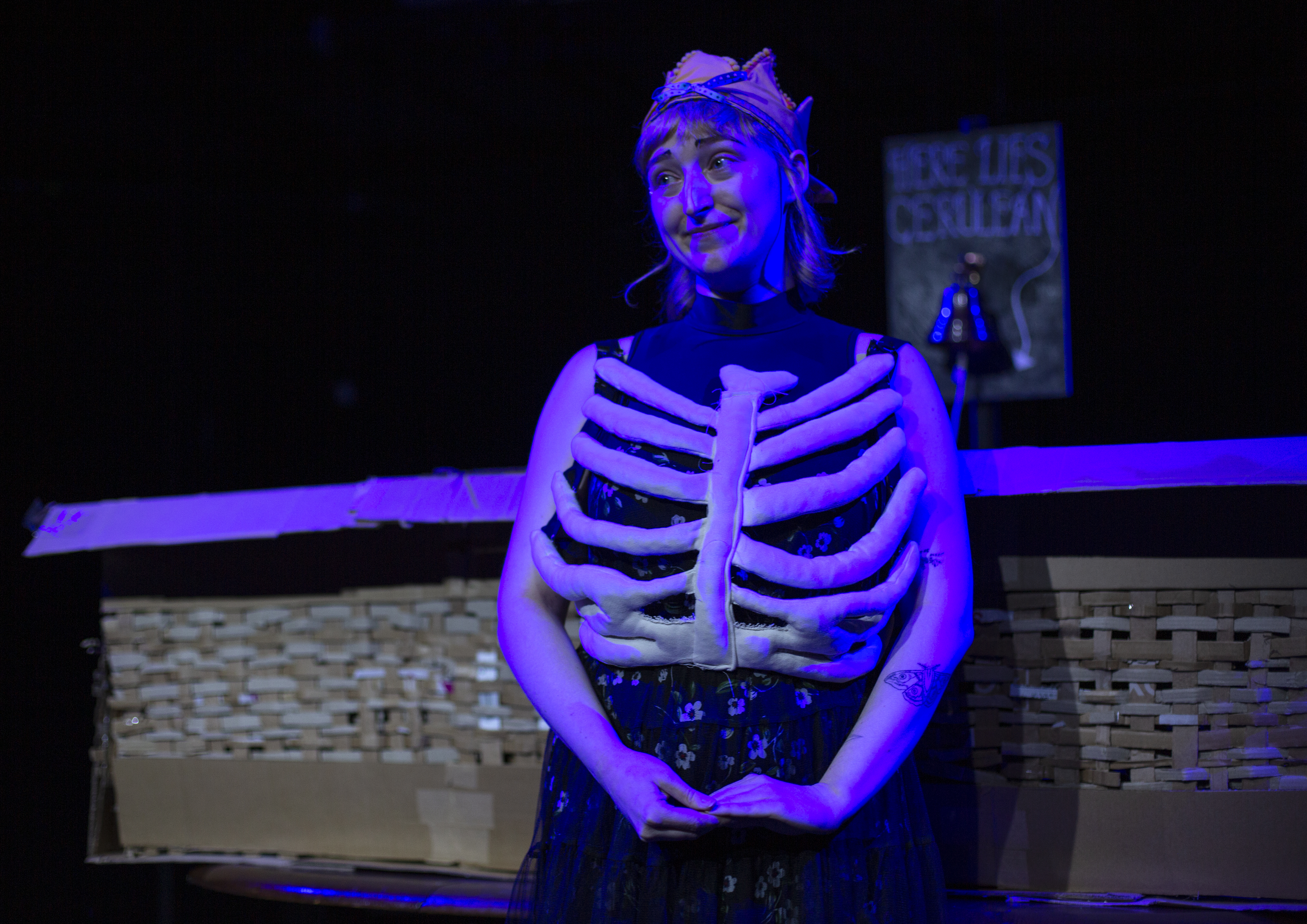 [no] good grief by Cerulean, Cerulean stands in front of a cardboard coffin with a fabric rib-cage tied around their body looking sweet but slightly cheeky